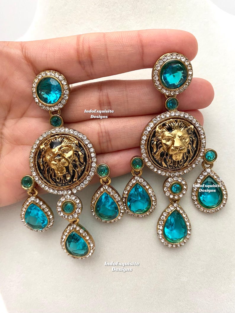 May include: A pair of gold tone earrings with a lion head design and turquoise blue crystal accents. The earrings feature a large round pendant with a lion head design, surrounded by smaller turquoise blue crystals. The lion head is gold tone and has a black enamel background. The earrings are suspended from a smaller round pendant with a turquoise blue crystal.