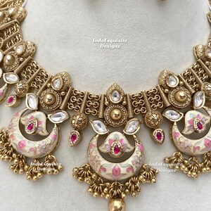 Pu&ograve; includere: Primo piano di un'ornata collana e orecchini color oro. La collana presenta pendenti a forma di mezzaluna color crema con disegni floreali rosa e accenti rossi. Pietre preziose chiare e rosse sono incastonate nel design.