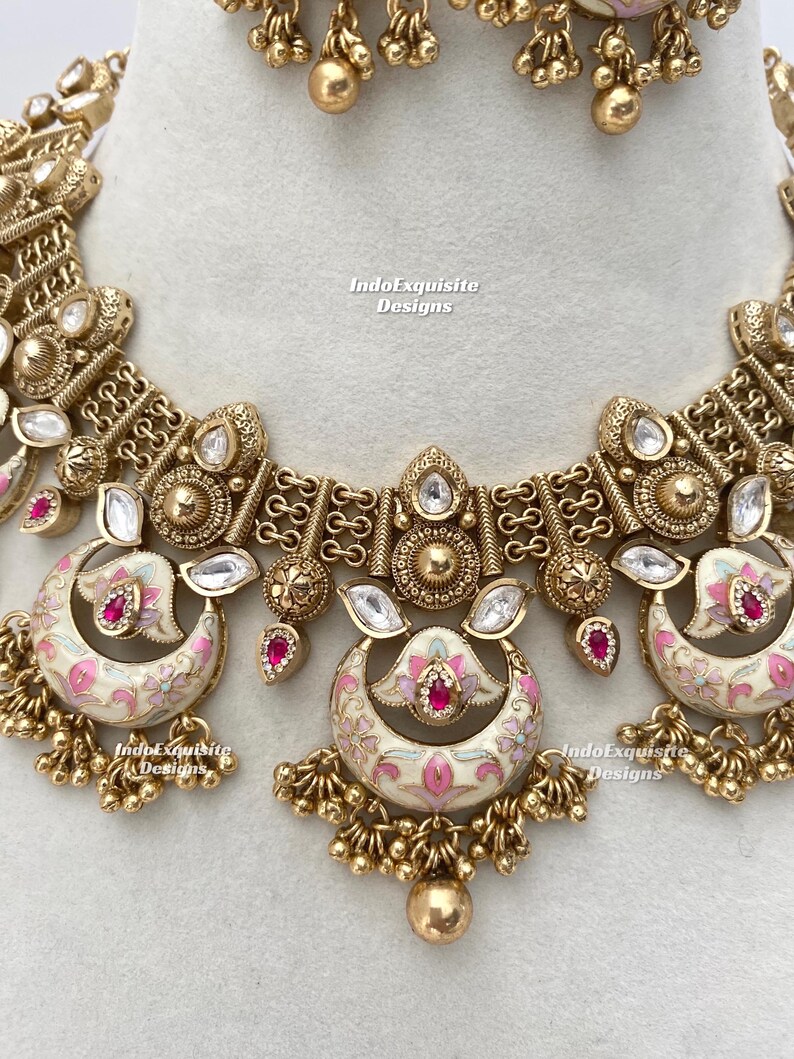 Pu&ograve; includere: Primo piano di un'ornata collana e orecchini color oro. La collana presenta pendenti a forma di mezzaluna color crema con disegni floreali rosa e accenti rossi. Pietre preziose chiare e rosse sono incastonate nel design.
