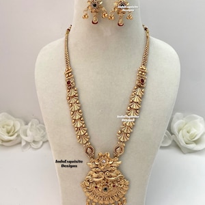 Può includere: Un set di collana e orecchini color oro, esposto su un manichino. La collana presenta un pendente dettagliato con accenti rossi e verdi, e gli orecchini hanno un design floreale con gocce rosse. Il set è ornato e probabilmente in metallo.