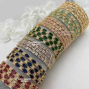 May include: A stack of gold bangles with different colored stones. The bangles are decorated with clear and colored square-shaped stones. The stones are set in gold metal. The bangles are stacked on a gray surface. The text "IndoExquisite Designs" is visible on the bottom bangle.