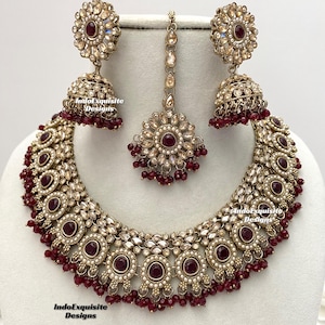 May include: A gold and maroon Indian bridal necklace set with a large pendant, two earrings, and a maang tikka. The necklace features a large round pendant with a maroon stone surrounded by smaller stones and pearls. The earrings are similar in design to the pendant, with a smaller maroon stone. The maang tikka is a small, ornate piece with a maroon stone in the center.