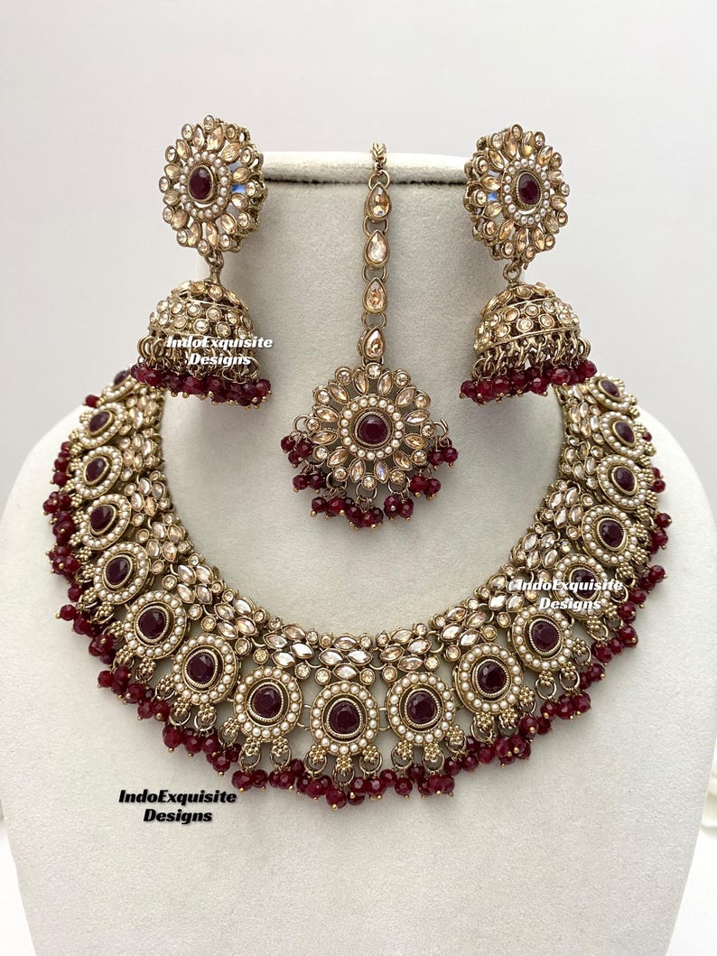 May include: A gold and maroon Indian bridal necklace set with a large pendant, two earrings, and a maang tikka. The necklace features a large round pendant with a maroon stone surrounded by smaller stones and pearls. The earrings are similar in design to the pendant, with a smaller maroon stone. The maang tikka is a small, ornate piece with a maroon stone in the center.