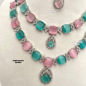 May include: A two-piece jewelry set featuring a silver necklace with alternating pink and green square-shaped stones and a matching pendant. The pendant is a teardrop shape with a green stone and silver accents. The necklace has a silver chain with a clasp.