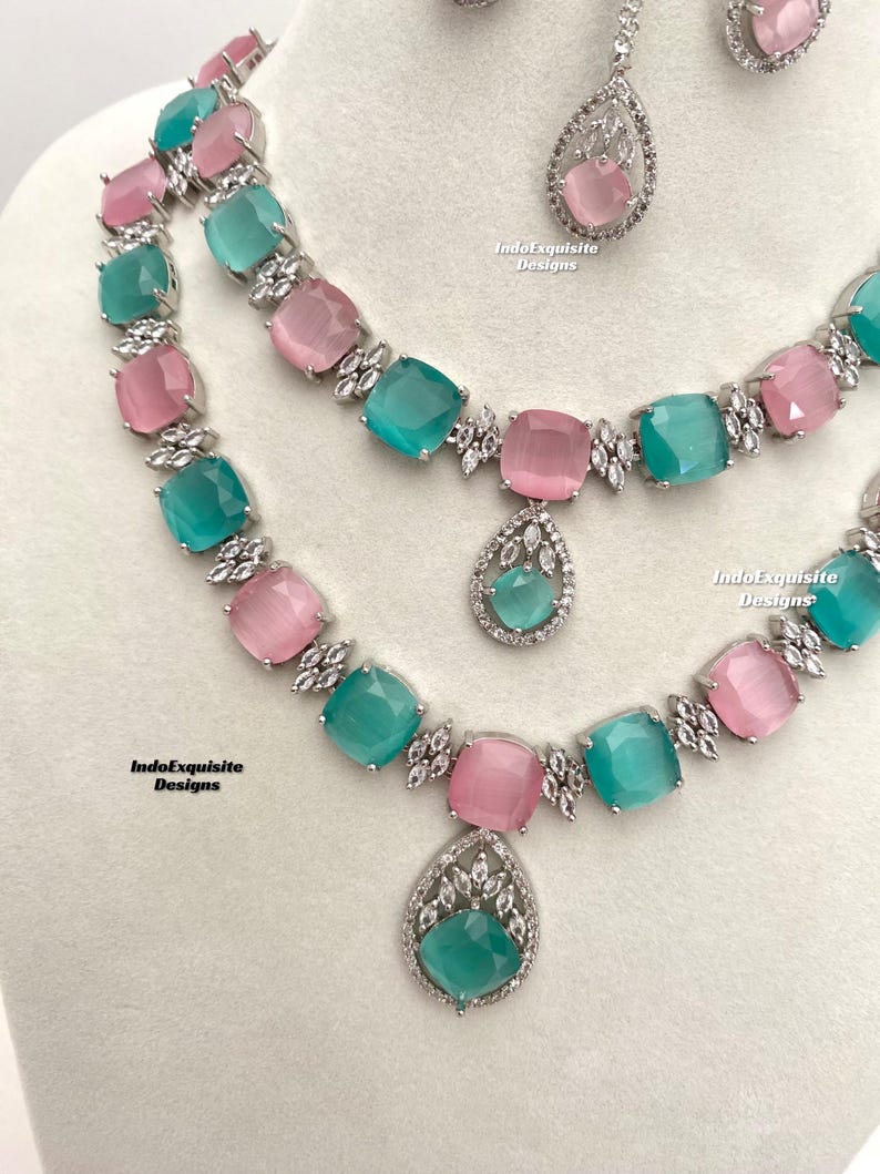 May include: A two-piece jewelry set featuring a silver necklace with alternating pink and green square-shaped stones and a matching pendant. The pendant is a teardrop shape with a green stone and silver accents. The necklace has a silver chain with a clasp.