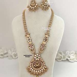 May include: A gold-toned necklace and earring set. The necklace features a pendant with a circular design, red and green accents, and small white stones. The earrings have a matching design. The text "IndoExquisite Designs" is visible.
