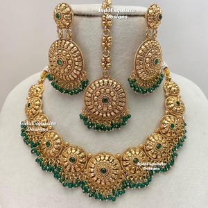 May include: A gold-coloured jewellery set comprising a necklace, earrings, and a headpiece. The set features a detailed, circular design with green accents. The necklace and earrings have dangling green beads. The headpiece has a central, matching circular design.