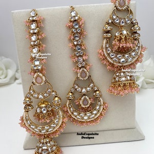 Puede incluir: Un conjunto de joyer&iacute;a en tono dorado que incluye dos pendientes largos y un adorno para la cabeza, decorados con piedras transparentes, peque&ntilde;as cuentas rosas y detalles de campana jhumka. Las piezas llevan el texto IndoExquisite Designs.