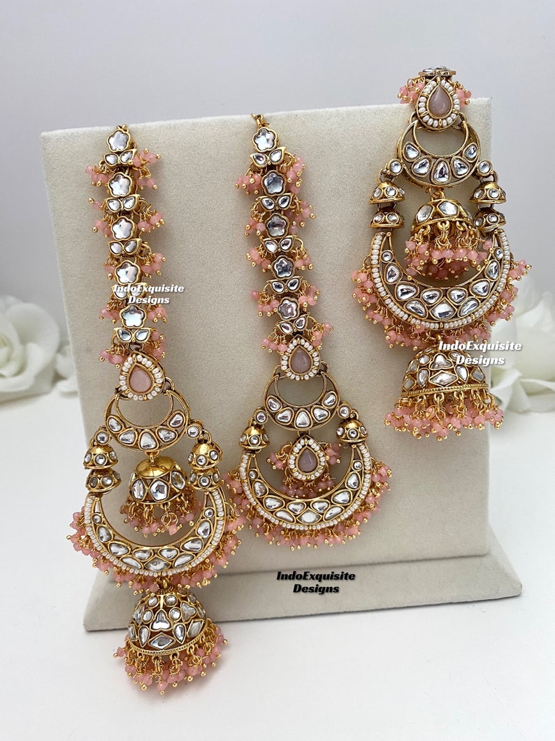 Puede incluir: Un conjunto de joyer&iacute;a en tono dorado que incluye dos pendientes largos y un adorno para la cabeza, decorados con piedras transparentes, peque&ntilde;as cuentas rosas y detalles de campana jhumka. Las piezas llevan el texto IndoExquisite Designs.