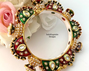 Puede incluir: Un brazalete dorado ornamentado con detalles de esmalte rojo, verde y blanco. El brazalete presenta un patrón repetitivo de formas de lágrima y está adornado con pequeñas perlas y gemas transparentes. El texto "IndoExquisite Designs" es visible.