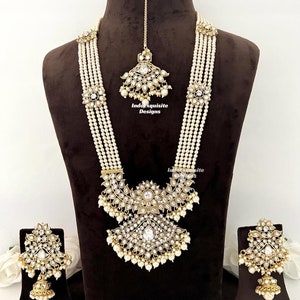 May include: An ornate, gold-toned jewelry set featuring a long necklace, earrings, and a headpiece. The necklace has multiple strands of white beads with floral and geometric accents. The earrings and headpiece mirror the necklace's design, with clear stones and dangling pearls.