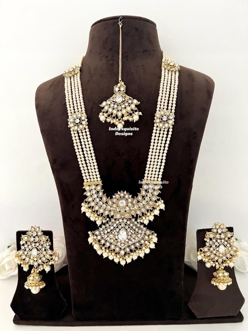May include: An ornate, gold-toned jewelry set featuring a long necklace, earrings, and a headpiece. The necklace has multiple strands of white beads with floral and geometric accents. The earrings and headpiece mirror the necklace's design, with clear stones and dangling pearls.