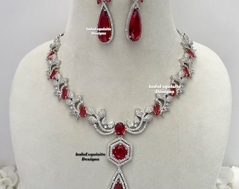 May include: A silver necklace and earring set with red gemstones. The necklace has a floral design with red accents and a large pendant. The earrings have a teardrop shape, mirroring the pendant. The text "IndoExquisite Designs" is visible on the jewelry.