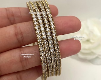 Puede incluir: Un conjunto de cuatro brazaletes dorados, cada uno con una hilera de piedras claras de corte redondo engastadas en un canal. El texto IndoExquisite Designs es visible en los brazaletes. La joyería tiene un acabado metálico y forma circular.