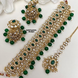Peut inclure: Un ensemble de bijoux doré comprenant un collier ras du cou, des boucles d'oreilles et un serre-tête. L'ensemble est orné de pierres précieuses vert émeraude et de petites perles blanches. Le collier et les boucles d'oreilles ont des perles vertes pendantes. Le texte "IndoExquisite Designs" est visible.