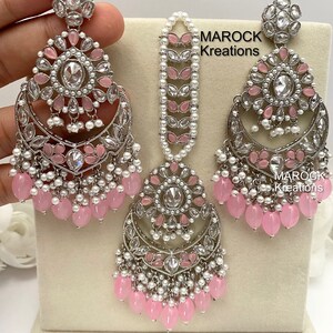 May include: A silver and pink bridal jewelry set with a teardrop-shaped pendant, earrings, and a maang tikka. The set features pink and white beads, clear stones, and intricate designs.