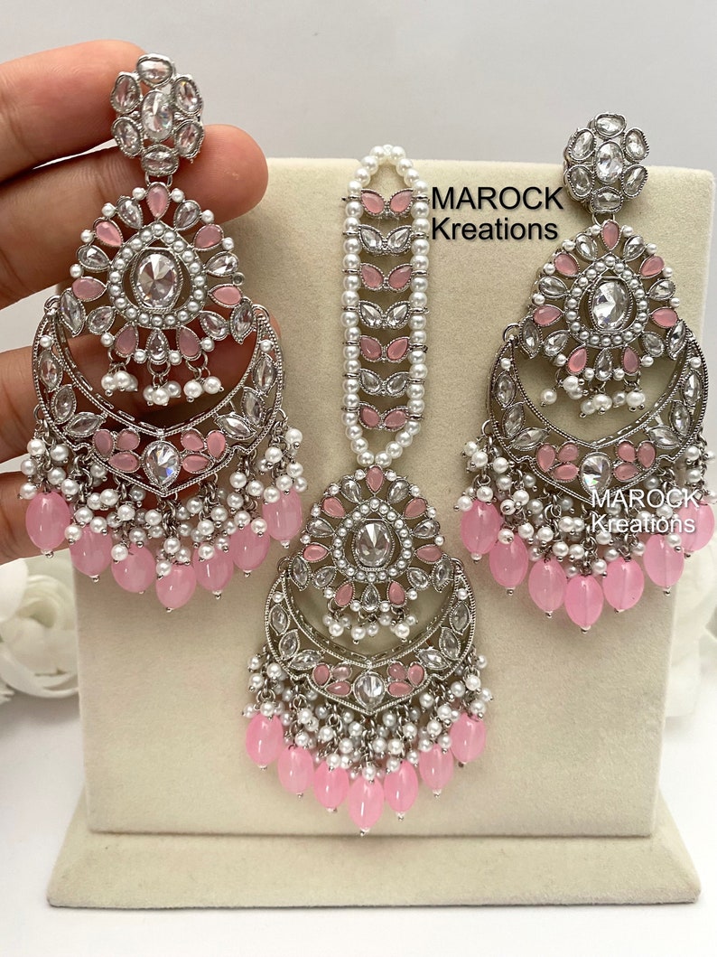 May include: A silver and pink bridal jewelry set with a teardrop-shaped pendant, earrings, and a maang tikka. The set features pink and white beads, clear stones, and intricate designs.