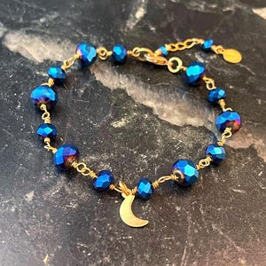 May include: A gold-toned bracelet featuring faceted blue beads and a small crescent moon charm. The beads are strung on gold wire, creating a delicate and elegant design. The bracelet has an adjustable chain.