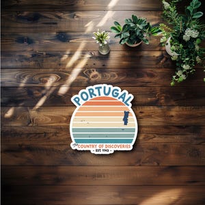 May include: A vintage-style sticker with a sunset graphic and the text "PORTUGAL" in blue letters. The sticker also includes the text "The Country of Discoveries - Est 1143 -" in a smaller font.