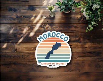 May include: A vintage-style sticker featuring a silhouette of Morocco with a sunset background. The sticker says "Morocco" in a retro font and "The Kingdom of the West - Est. 1956 -" in a smaller font.