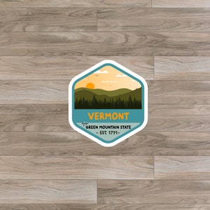 May include: A blue and white hexagonal sticker with a sunset scene over a green mountain range. The text "The Green Mountain State - Est. 1791 -" is written in white on a blue banner below the mountains. The text "Vermont" is written in white on a blue banner above the mountains.