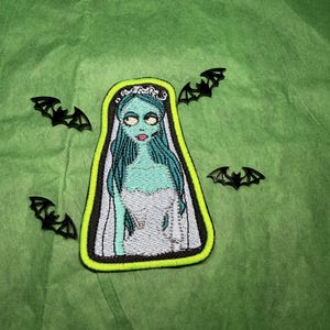 May include: An embroidered patch featuring a character with pale blue skin, long blue hair, and a white dress, surrounded by a bright neon green border. Four small, black, flat, plastic bat-shaped decorations are arranged around the patch.