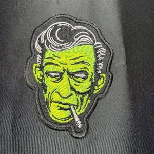 May include: An embroidered patch featuring a stylized illustration of a person with bright green skin, dark hair, and a cigarette in their mouth. The design uses black outlines and white highlights, finished with a thick black border on a black fabric.