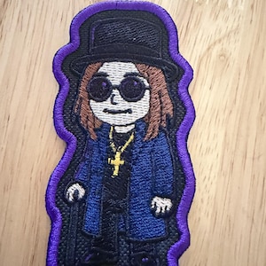 Puede incluir: Un parche bordado que muestra una figura de dibujos animados con un sombrero de copa negro, gafas de sol redondas oscuras y un abrigo largo azul sobre una camisa negra. La figura tiene cabello castaño, un collar con cruz dorada y un bastón negro, todo bordeado en morado.