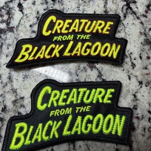 May include: Two embroidered patches featuring the text "CREATURE FROM THE BLACK LAGOON" in a stylized font. The top patch has yellow lettering with a thin orange outline, while the bottom patch features bright lime green lettering on black fabric.