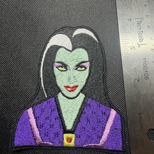 May include: An embroidered patch featuring a character with pale green skin, black hair with a white streak, pink eyeshadow, and red lips, wearing a purple garment with a yellow square accent. A metal ruler shows the patch is 3.5 inches tall.