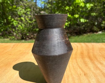 Puede incluir: Un jarrón de madera artesanal marrón oscuro con una forma geométrica única. El jarrón tiene una parte superior ancha y acampanada y una base cónica, creando una silueta llamativa. La veta de la madera es visible, agregando textura. El jarrón mide aproximadamente 15 cm de alto.