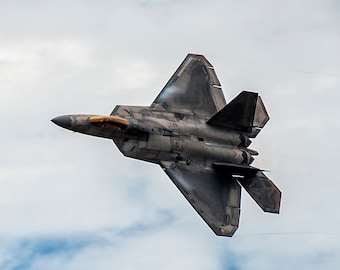May include: A gray and brown fighter jet flies in a cloudy sky. The jet is flying in a steep bank, with its wings tilted upwards. The jet is leaving a trail of smoke behind it.