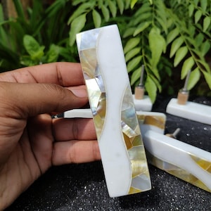 May include: A set of four white and gold cabinet door pulls made of mother of pearl. The pulls have a wavy design and are held in a hand.