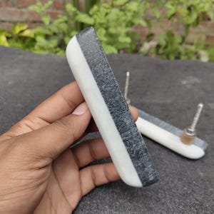 May include: A pair of rectangular cabinet pulls with a contrasting design. The pulls feature a combination of white, gray, and black marble. Each pull has a silver-toned metal stem for mounting. The pulls are designed for furniture.