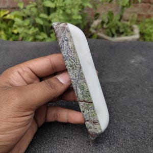 May include: A rectangular, polished stone with a white, smooth surface on one side and a textured, green and brown pattern on the other. The stone is held in a hand, showcasing its contrasting textures and colors.