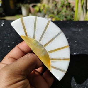 May include: A white and gold half-circle shaped decorative object with a sunburst design. The object is made of mother of pearl and has a gold trim.