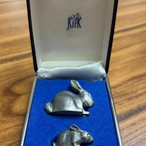 May include: A set of two silver-toned metal rabbit figurines in a presentation box. The box has a blue velvet interior and the brand name "Kirk" is printed on the inside lid. The rabbits are detailed with textured fur.