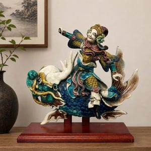 May include: A ceramic figurine depicting a figure riding a fish. The figure is dressed in colorful robes, with a smiling face and outstretched arm. The fish is blue and white, mounted on a wooden base. The figurine is a decorative item.