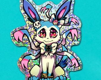 以下が含まれることがあります： 青い耳とピンクの耳、白い体、ピンクのリボンをつけた漫画のキャラクターが描かれたキラキラしたステッカー。キャラクターは大きな目と頭蓋骨を持っています。
