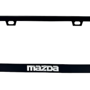 Puede incluir: Marco negro para matr&iacute;cula con el logotipo de Mazda en plata. El marco rectangular tiene esquinas redondeadas y orificios de montaje en la parte superior. El logotipo de Mazda est&aacute; centrado en la parte inferior.