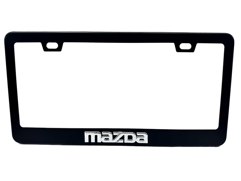 Puede incluir: Marco negro para matr&iacute;cula con el logotipo de Mazda en plata. El marco rectangular tiene esquinas redondeadas y orificios de montaje en la parte superior. El logotipo de Mazda est&aacute; centrado en la parte inferior.