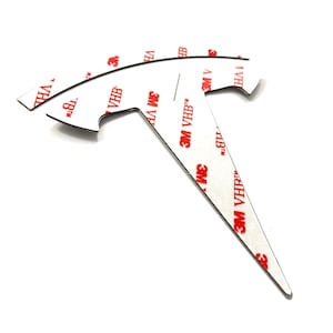 K&ouml;nnte beinhalten: Ein schwarzes Metallemblema in T-Form mit einer spitzen Spitze. Das Emblem hat eine wei&szlig;e Klebeunterseite mit dem in Rot gedruckten Text "3M VHB".