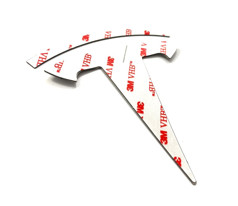 K&ouml;nnte beinhalten: Ein schwarzes Metallemblema in T-Form mit einer spitzen Spitze. Das Emblem hat eine wei&szlig;e Klebeunterseite mit dem in Rot gedruckten Text "3M VHB".