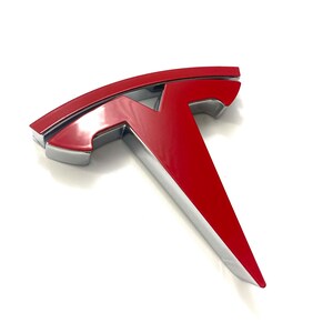 K&ouml;nnte beinhalten: Ein rotes und silbernes Tesla-Logo. Das Logo ist ein stilisiertes T mit einer abgerundeten Oberseite und einer spitzen Unterseite.