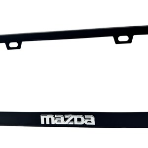 Puede incluir: Marco negro para matr&iacute;cula con el logotipo de Mazda en plata. El marco tiene forma rectangular con esquinas redondeadas y dos orificios de montaje en la parte superior. El logotipo de Mazda est&aacute; centrado en la parte inferior.