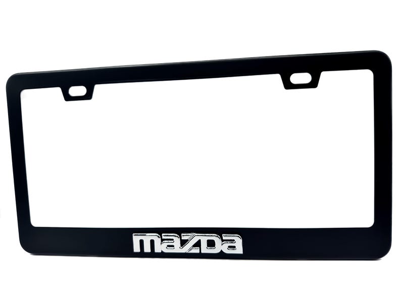 Puede incluir: Marco negro para matr&iacute;cula con el logotipo de Mazda en plata. El marco tiene forma rectangular con esquinas redondeadas y dos orificios de montaje en la parte superior. El logotipo de Mazda est&aacute; centrado en la parte inferior.