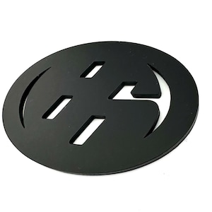 May include: A black, oval-shaped emblem with a stylized design. The emblem features a series of rectangular cutouts and curved edges, creating a unique pattern. The design is set against a white background, highlighting the black color.