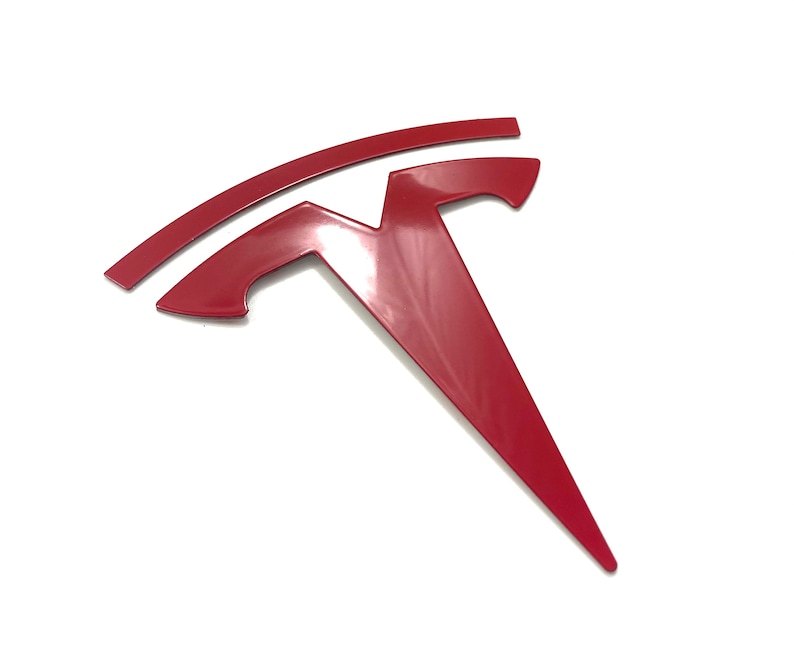 K&ouml;nnte beinhalten: Ein rotes Tesla-Logo aus Metall mit einem stilisierten 'T' und einer geschwungenen Linie dar&uuml;ber.