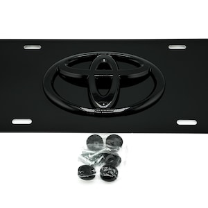 May include: Black license plate frame with a raised, black Toyota logo. The frame is made of metal and has four mounting holes.