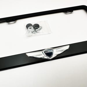 May include: Black metal license plate frame with a silver Genesis logo and wings. The frame comes with four black plastic mounting screws.